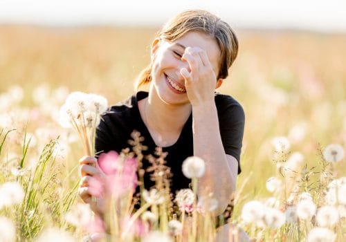 Pretty girl teenager sitting in field with dandelions and flowers, looking at camera and smiling outdoors. Beautiful young female person at nature in sunny day portrait