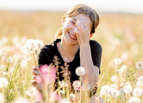 Pretty girl teenager sitting in field with dandelions and flowers, looking at camera and smiling outdoors. Beautiful young female person at nature in sunny day portrait